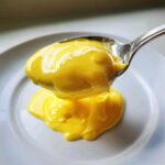 A spoonful of thick, bright yellow hollandaise sauce being lifted above a small dollop on a white plate.