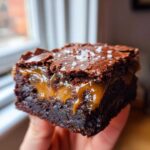 Close-up of a fudgy caramel brownie slice oozing with gooey caramel and topped with flaky sea salt.