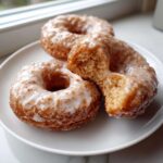 Three glazed cake donuts on a white plate, with one donut broken in half showing the fluffy interior.