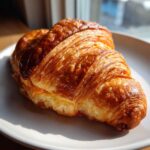 A close-up of a golden brown baked ham and cheese croissant resting on a light plate.