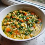 Close-up of a white bowl filled with vibrant greek chickpea soup, featuring chickpeas, carrots, orzo pasta, and fresh parsley.