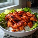 Close-up of a white bowl filled with white rice topped generously with bright red, glazed harissa chicken and fresh green lettuce.