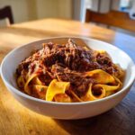 A close-up of a white bowl filled with wide pappardelle pasta topped generously with rich, shredded beef ragu.