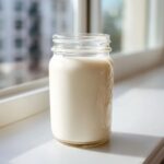 A glass mason jar filled with creamy homemade coffee creamer sitting on a bright windowsill.