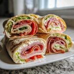 Four stacked cross-sections of Italian pinwheels showing layers of deli meat, cheese spread, lettuce, and tomato in a tortilla.