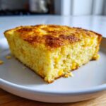 A close-up of a golden-brown slice of moist jiffy corn casserole featuring visible corn kernels.