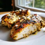 Close-up of a sliced, juicy greek chicken breast seasoned with herbs and topped with crumbled feta cheese.