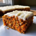 Close-up of a moist slice of carrot cake bars showing shredded carrots and nuts, topped with thick cream cheese frosting.