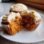 Close-up of a moist pumpkin spice cupcake cut in half showing the orange crumb and cream cheese frosting.