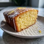 Close-up of a moist slice of lemon olive oil cake with white glaze and zest topping on a white plate.