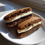 Two homemade oatmeal cream pies with dark brown cookies and thick white cream filling, resting on a white plate.