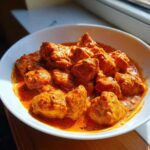 A close-up, appetizing shot of tender chicken pieces swimming in a vibrant, orange-red sauce, showcasing the rich texture of butter chicken.