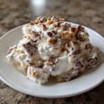 A close-up of a serving of creamy Snickers salad mixed with chopped candy bars and nuts on a white plate.