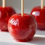 A close-up of a perfectly coated, shiny red candy apple with visible gold glitter specks, ready to eat.