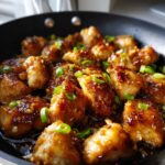Close-up of glazed honey garlic chicken pieces sizzling in a black skillet, topped with sesame seeds and sliced green onions.