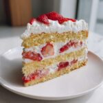 A tall, moist slice of strawberry shortcake cake with layers of sponge cake, whipped cream, and fresh strawberries.