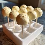 A display of smooth, white chocolate cake pops topped with sprinkles, standing upright in a white holder.