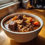 A close-up of a steaming white bowl filled with rich beef barley soup, featuring chunks of beef, carrots, and celery.