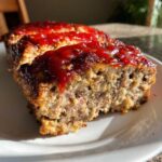 Close-up of a thick slice of stove top stuffing meatloaf topped with a shiny red ketchup glaze, served on a white plate.