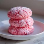Three bright pink strawberry cake mix cookies stacked on a white plate, dusted with powdered sugar.