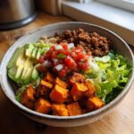 A vibrant sweet potato taco bowl featuring seasoned sweet potato cubes, ground meat, avocado, lettuce, and fresh salsa.