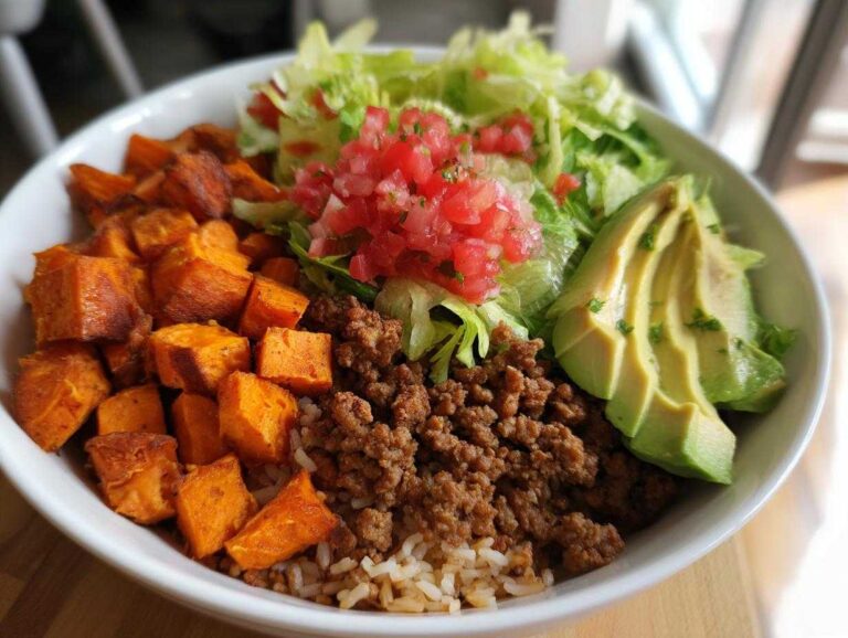 A close-up of a sweet potato taco bowl featuring roasted sweet potatoes, ground meat, rice, lettuce, pico de gallo, and sliced avocado.