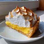 A tall, perfect slice of lemon meringue pie showing a thick layer of bright yellow curd and high, toasted meringue peaks.