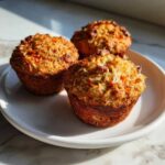 Three freshly baked morning glory muffins topped with shredded coconut and pecans, sitting on a white plate.