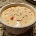 Close-up of a creamy, warm cheese dip mixed with diced red tomatoes and sprinkled with green herbs.