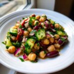 Close-up of a vibrant, dressed dense bean salad featuring chickpeas, cucumber, red onion, and kidney beans on a white plate.