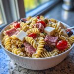Close-up of a white bowl filled with italian pasta salad featuring rotini, cubed cheese, salami, and cherry tomatoes.