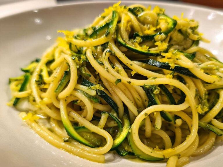 A close-up of a bowl of zucchini pasta mixed with spaghetti, topped with bright lemon zest.