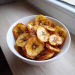 Close-up of a white bowl filled with golden, crispy air fryer banana chips dusted with cinnamon.