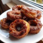 A close-up of several golden brown air fryer cinnamon apple rings coated in sugar and spice on a white plate.