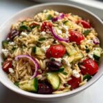 A close-up view of a vibrant greek orzo pasta salad mixed with cherry tomatoes, cucumbers, feta, and red onion.