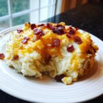 A serving of creamy mashed potato casserole topped with melted cheddar cheese and crispy bacon bits on a white plate.