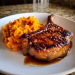 A perfectly cooked, glazed apple cider pork chop served next to a mound of bright orange mashed sweet potatoes.