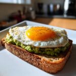 Close-up of a perfectly cooked avocado egg sandwich featuring mashed avocado on whole wheat toast topped with a sunny-side-up egg.
