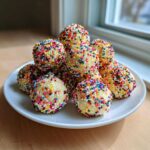 A pile of bright, round birthday cake batter protein balls completely covered in rainbow sprinkles, served on a white plate.