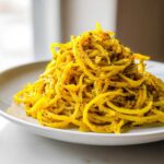 A close-up of bright yellow black pepper turmeric cauliflower and garlic noodles piled high on a white plate.