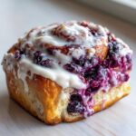 A close-up of one fluffy, square blueberry cinnamon rolls topped with thick white icing and bursting with purple blueberry filling.
