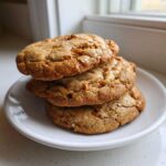 Three chewy brown butter coffee toffee cookies stacked on a small white plate near a bright window.