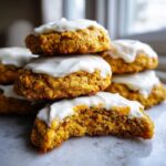 Stack of soft carrot cake cookies recipe treats topped with thick white cream cheese frosting, one cookie is bitten.