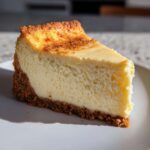 A thick, creamy slice of cheesecake recipe on a white plate, featuring a graham cracker crust and a lightly browned top.
