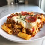 A close-up of a serving of cheesy pizza pasta bake topped with melted mozzarella, pepperoni slices, and herbs.