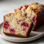 Close-up of a moist slice of cherry almond poppyseed bread showing bright red fruit and black poppy seeds throughout the crumb.