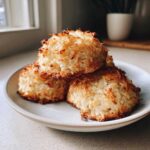 Three golden-brown, textured chewy coconut cookies stacked on a light gray plate near a window.