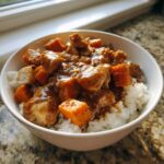 A white bowl filled with white rice topped with chunks of chicken and sweet potato covered in a savory brown sauce, making a delicious chicken sweet potato rice bowl.
