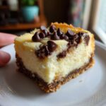 A decadent square of chocolate chip cheesecake bars with a thick graham cracker crust, topped with melted and whole chocolate chips.