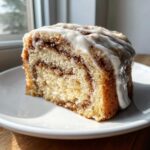 A close-up of a moist slice of cinnamon roll cake recipe with a visible swirl of cinnamon filling and thick white icing.